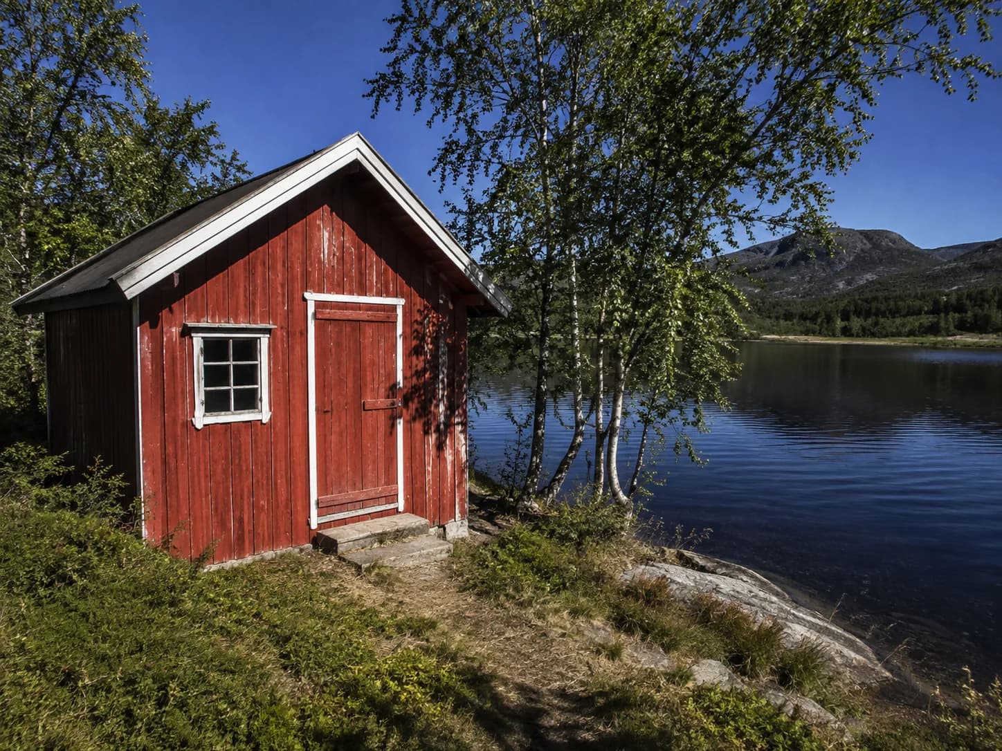 Rød trehytte ved et vann i hardt sollys, med skarpe skygger og høy kontrast.